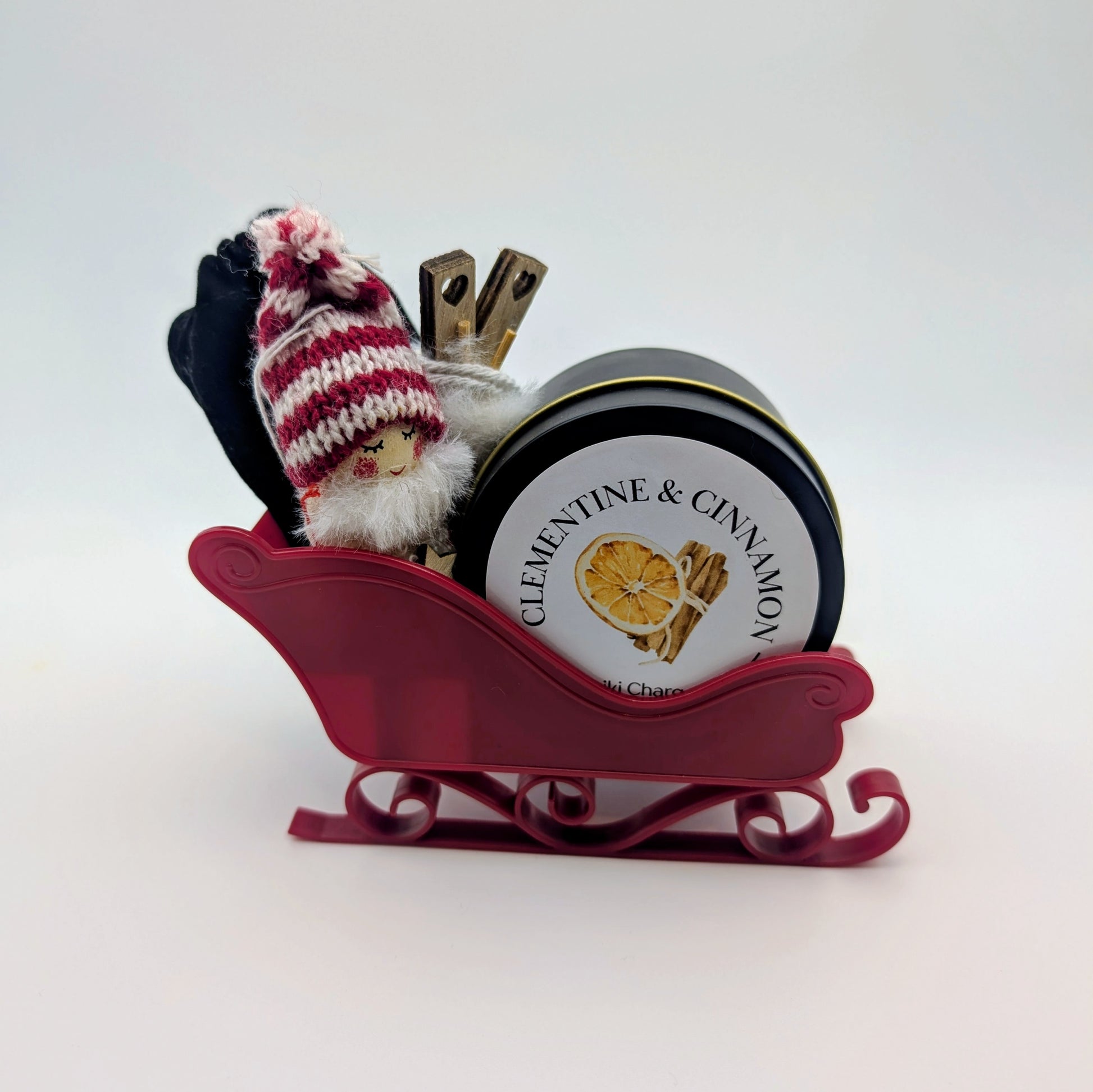 Red sleigh with a small Santa figure and a cylindrical container labeled 'Clementine & Cinnamon' on a white background.